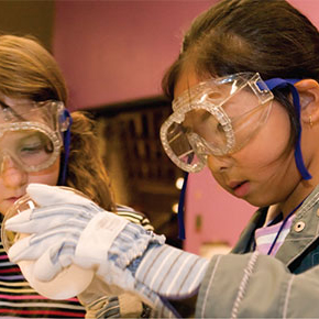 Two children with safety glasses and gloves working on a science experiment