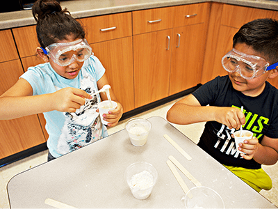 Two children wear safety goggles while performing a science experiment