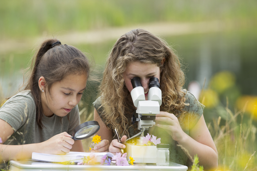Two students are outside examining nature using a microscope and a magnifying glass