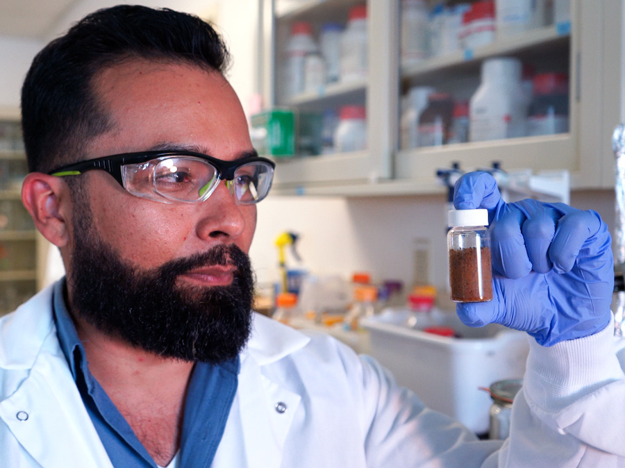 Javier wearing safety glasses, a lab coat and gloves, holding up a vial